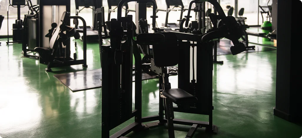 Silhouetten van fitnessapparatuur in een slecht verlichte fitnessruimte met een groene vloer. Het ochtendlicht valt door grote ramen naar binnen en creëert een rustige sfeer.